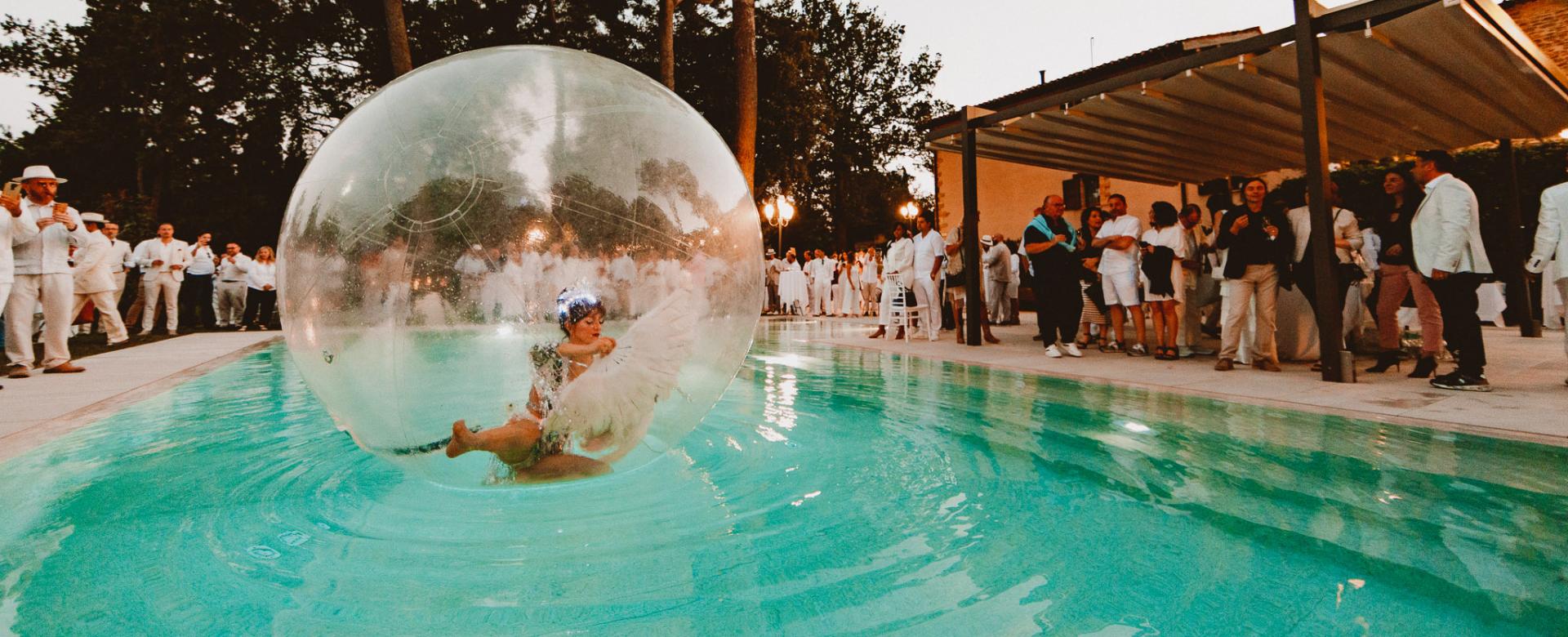Esibizione in piscina con una persona dentro una sfera trasparente, pubblico vestito di bianco.