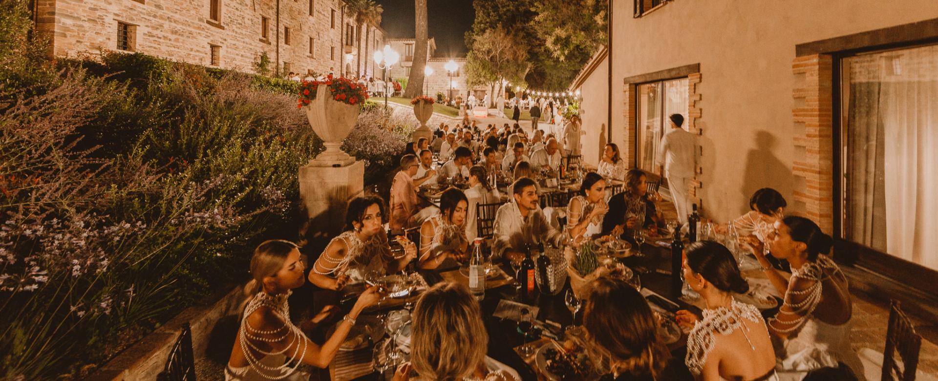 Cena all'aperto in un ambiente rustico, con molte persone che indossano abiti eleganti.